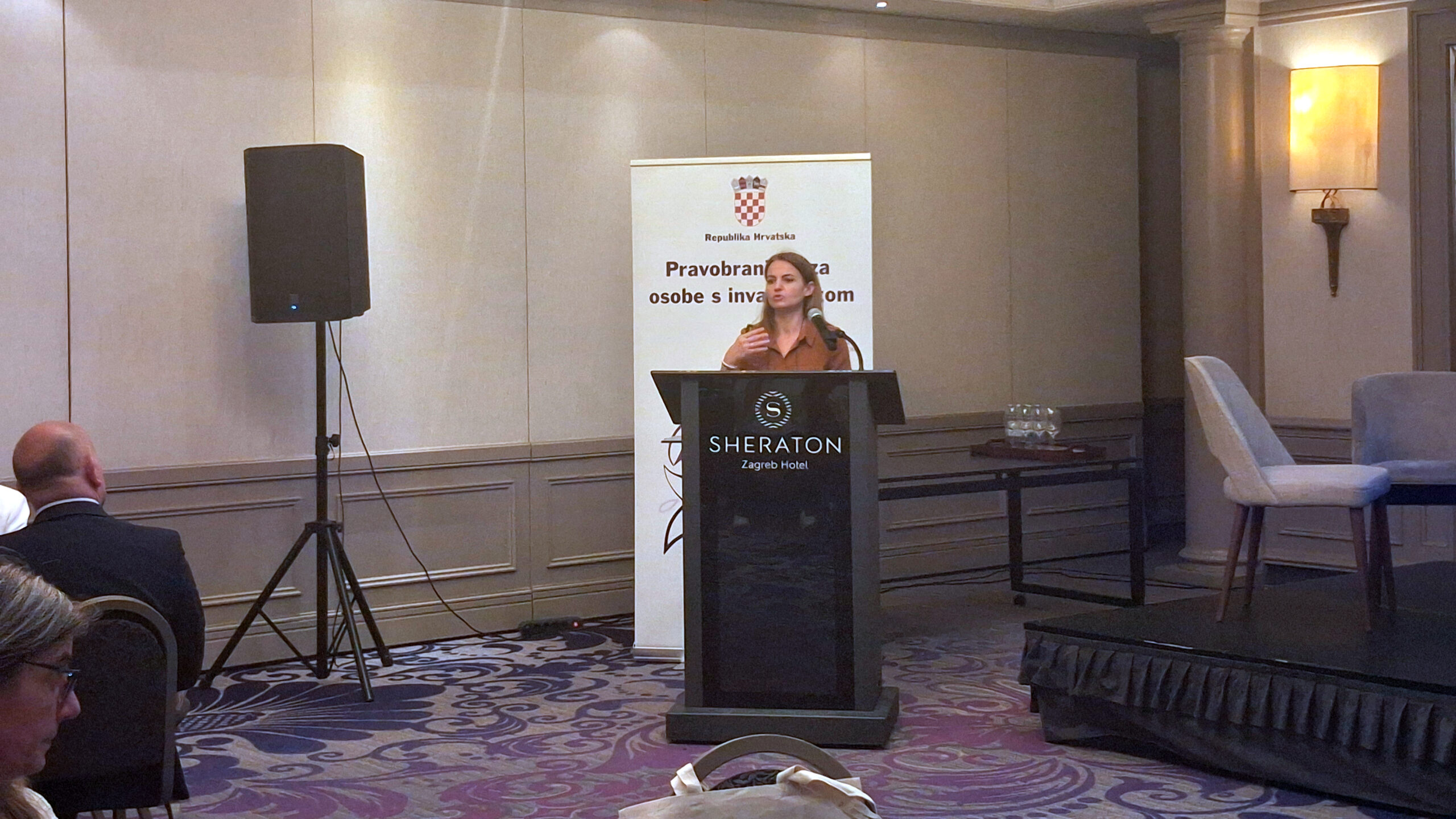 Žena govori za govornicom na konferenciji, ispred bannera Pravobranitelja za osobe s invaliditetom u hotelu Sheraton.