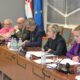 Na fotografiji je prikazan sastanak u konferencijskoj dvorani Hrvatskoga sabora. Za dugim drvenim stolom sjedi nekoliko sudionika rasprave, među kojima su zastupnici i predstavnici institucija. U prvom planu vidi se žena kratke plave kose koja govori u mikrofon, dok ostali sudionici pažljivo slušaju. Na stolu se nalaze dokumenti, mikrofoni, slušalice i boce vode. U pozadini su istaknute zastave Hrvatske i Europske unije, što upućuje na službeni i međunarodni karakter sastanka. Atmosfera djeluje formalno i radno, a sudionici su usredotočeni na raspravu o temama vezanim uz prava i uključivanje osoba s invaliditetom u politički život.
