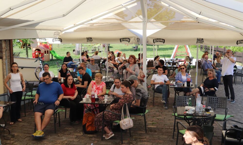 Na fotografiji je prikazano okupljanje većeg broja ljudi na natkrivenoj terasi ugostiteljskog objekta tijekom sunčanog dana. Posjetitelji sjede za stolovima, razgovaraju i prate događaj koji se odvija izvan kadra, prema lijevoj strani fotografije. Atmosfera djeluje opušteno i društvena. Ispod velikih bijelih tendi nalaze se stolovi s pićem, kavom i bocama vode, a na pojedinim stolovima vide se i osobni predmeti poput torbi. U prvom planu sjedi nekoliko skupina ljudi različite dobi, dok se u pozadini vidi još gostiju raspoređenih po terasi. Izvan terase nalazi se travnata površina s dječjim igralištem, uključujući mali tobogan i druge sprave za igru, što upućuje na obiteljski ambijent. Na suncobranima se nalazi logo pivskog brenda, a cijeli prostor odiše ugođajem ljetnog druženja na otvorenom. Fotografija ostavlja dojam zajedništva i neformalnog okupljanja, poput predstavljanja, predavanja, glazbenog nastupa ili drugog društvenog događaja kojem prisutni posvećuju pažnju.