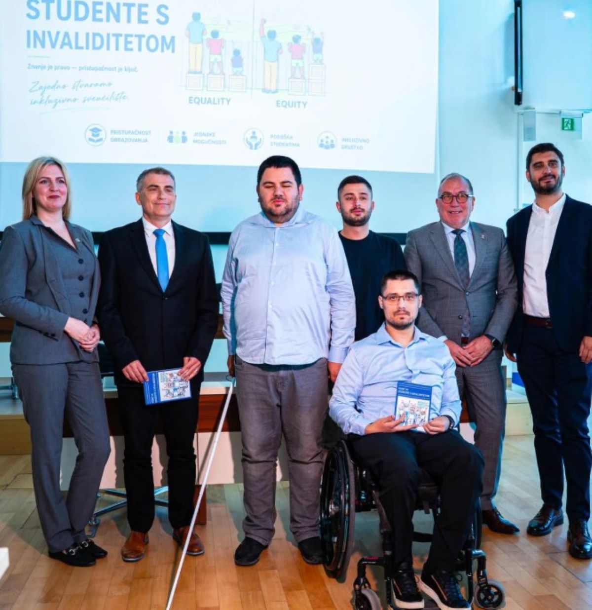 Na fotografiji se nalazi skupina ljudi okupljena na predstavljanju vodiča za studente s invaliditetom. U prvom planu pozira šest osoba, među kojima je jedan muškarac u invalidskim kolicima koji drži primjerak vodiča. Ostali sudionici stoje uz njega, odjeveni uglavnom u poslovnu i svečanu odjeću. U pozadini je projekcija s natpisom o studentima s invaliditetom te ilustracijama koje prikazuju jednakost i pravednost (“Equality” i “Equity”). Na ekranu se također vide pojmovi poput pristupačnosti obrazovanja, jednakih mogućnosti, podrške studentima i inkluzivnog društva. Atmosfera djeluje službeno i edukativno, kao dio konferencije ili javnog predstavljanja projekta vezanog uz inkluzivno obrazovanje.