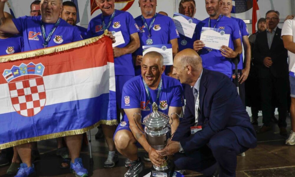 Na fotografiji je prikazan svečani trenutak dodjele pehara hrvatskoj reprezentaciji osoba s invaliditetom nakon osvajanja naslova viceprvaka svijeta u sportskom ribolovu. U prvom planu nekoliko članova reprezentacije, odjevenih u plave sportske majice s hrvatskim grbom, ponosno drže veliki srebrni pehar i hrvatsku zastavu. Natjecatelji oko vrata imaju medalje, a neki u rukama drže diplome i priznanja. Atmosfera je svečana i emotivna, a osmijesi i slavlje članova ekipe pokazuju zadovoljstvo velikim sportskim uspjehom. U pozadini se vide ostali sudionici i uzvanici na pozornici tijekom završne ceremonije prvenstva u Ivancu.