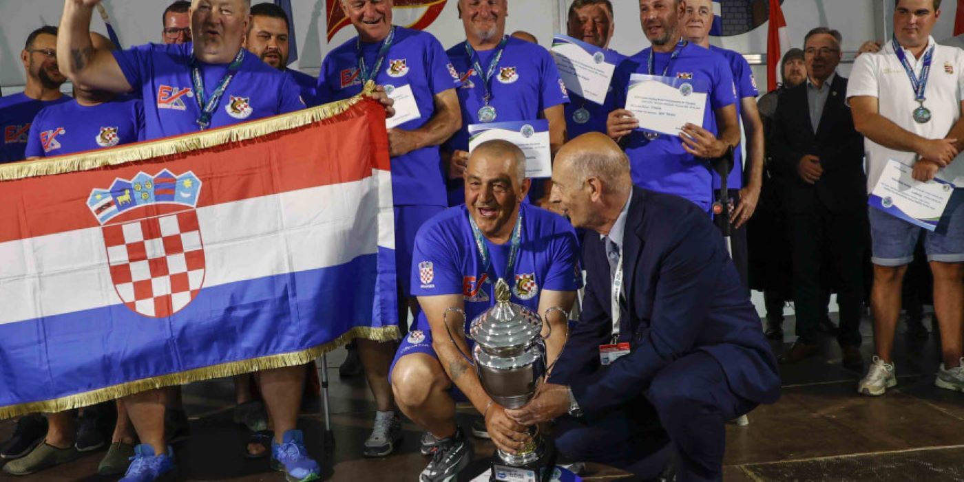Na fotografiji je prikazan svečani trenutak dodjele pehara hrvatskoj reprezentaciji osoba s invaliditetom nakon osvajanja naslova viceprvaka svijeta u sportskom ribolovu. U prvom planu nekoliko članova reprezentacije, odjevenih u plave sportske majice s hrvatskim grbom, ponosno drže veliki srebrni pehar i hrvatsku zastavu. Natjecatelji oko vrata imaju medalje, a neki u rukama drže diplome i priznanja. Atmosfera je svečana i emotivna, a osmijesi i slavlje članova ekipe pokazuju zadovoljstvo velikim sportskim uspjehom. U pozadini se vide ostali sudionici i uzvanici na pozornici tijekom završne ceremonije prvenstva u Ivancu.
