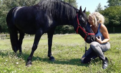 Žena kleči na travi i nježno prislanja glavu uz crnog konja na livadi.