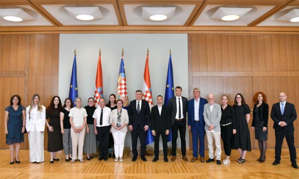 Na fotografiji je snimljena skupina ljudi tijekom službenog susreta u reprezentativnoj dvorani s drvenim zidnim oblogama. U sredini stoji predsjednik Republike, okružen predstavnicima Hrvatskog saveza gluhoslijepih osoba Dodir i drugim sudionicima sastanka. Iza okupljenih nalaze se zastave Republike Hrvatske i Europske unije, što naglašava službeni karakter događaja. Sudionici su poredani u jednom redu i poziraju za zajedničku fotografiju. Većina je odjevena u poslovnu ili svečanu odjeću, a svi gledaju prema kameri s ozbiljnim ili blagim osmijehom. Fotografija prikazuje završetak ili dio protokolarnog susreta održanog povodom obilježavanja Međunarodnog dana gluhosljepoće.