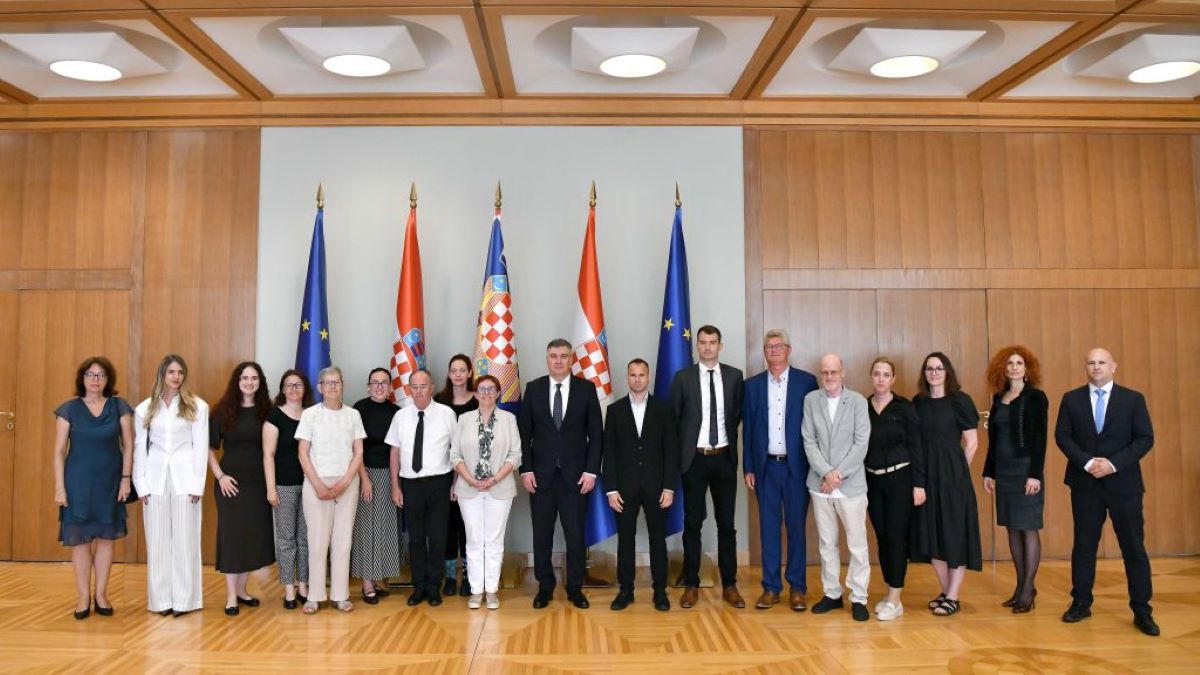 Na fotografiji je snimljena skupina ljudi tijekom službenog susreta u reprezentativnoj dvorani s drvenim zidnim oblogama. U sredini stoji predsjednik Republike, okružen predstavnicima Hrvatskog saveza gluhoslijepih osoba Dodir i drugim sudionicima sastanka. Iza okupljenih nalaze se zastave Republike Hrvatske i Europske unije, što naglašava službeni karakter događaja. Sudionici su poredani u jednom redu i poziraju za zajedničku fotografiju. Većina je odjevena u poslovnu ili svečanu odjeću, a svi gledaju prema kameri s ozbiljnim ili blagim osmijehom. Fotografija prikazuje završetak ili dio protokolarnog susreta održanog povodom obilježavanja Međunarodnog dana gluhosljepoće.