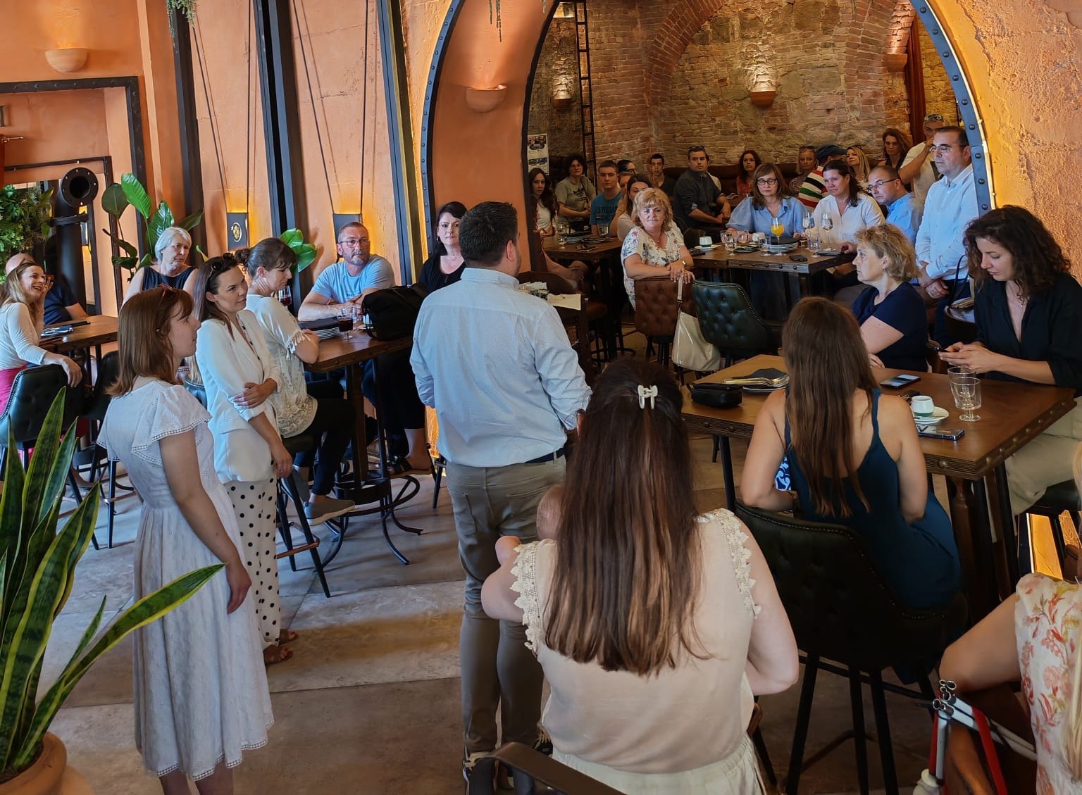 Na fotografiji je prikazan skup sudionika okupljenih na predstavljanju ili konferencijskom događanju u ugodnom unutarnjem prostoru rustikalnog uređenja. Prostor karakteriziraju kameni zidovi, lučni prolazi i topla rasvjeta koja stvara ugodnu atmosferu. U središtu fotografije nalazi se muškarac koji stoji okrenut prema okupljenima i obraća se prisutnima. Sudionici sjede za stolovima ili stoje uz zidove te pažljivo slušaju izlaganje. Na stolovima se mogu vidjeti čaše, šalice i promotivni materijali, što upućuje na poslovni ili društveni događaj. Okupljeni čine raznoliku skupinu žena i muškaraca različite dobi, a atmosfera djeluje profesionalno, ali istovremeno opušteno i prijateljski. U prvom planu nalaze se biljke koje dodatno doprinose ugodnom ambijentu prostora. Fotografija bilježi trenutak predstavljanja projekta ili službenog obraćanja, pri čemu je naglasak na interakciji među sudionicima, razmjeni informacija i zajedničkom sudjelovanju u događanju.