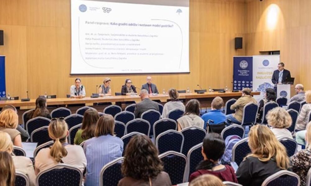 Na fotografiji je prikazan stručni skup u velikoj konferencijskoj dvorani. U prvom planu vide se sudionici koji sjede u redovima plavih stolica i prate panel-raspravu. Na pozornici za dugim stolom sjedi nekoliko panelista, dok jedan govornik stoji za govornicom s desne strane dvorane i obraća se okupljenima. Iza sudionika na pozornici nalazi se veliko projekcijsko platno s naslovom panel-rasprave „Kako graditi održiv i sustavan model podrške?“, a uz pozornicu su postavljeni promotivni roll-up plakati Sveučilišta u Zagrebu i skupa posvećenog inkluziji i različitosti. Prostor djeluje formalno i akademski, a atmosfera odražava stručni i edukativni karakter događanja.
