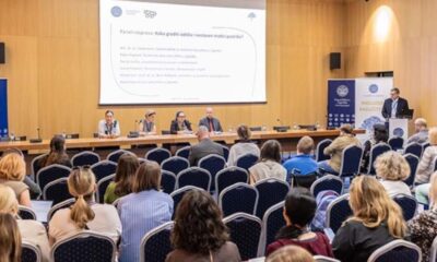 Na fotografiji je prikazan stručni skup u velikoj konferencijskoj dvorani. U prvom planu vide se sudionici koji sjede u redovima plavih stolica i prate panel-raspravu. Na pozornici za dugim stolom sjedi nekoliko panelista, dok jedan govornik stoji za govornicom s desne strane dvorane i obraća se okupljenima. Iza sudionika na pozornici nalazi se veliko projekcijsko platno s naslovom panel-rasprave „Kako graditi održiv i sustavan model podrške?“, a uz pozornicu su postavljeni promotivni roll-up plakati Sveučilišta u Zagrebu i skupa posvećenog inkluziji i različitosti. Prostor djeluje formalno i akademski, a atmosfera odražava stručni i edukativni karakter događanja.