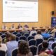 Na fotografiji je prikazan stručni skup u velikoj konferencijskoj dvorani. U prvom planu vide se sudionici koji sjede u redovima plavih stolica i prate panel-raspravu. Na pozornici za dugim stolom sjedi nekoliko panelista, dok jedan govornik stoji za govornicom s desne strane dvorane i obraća se okupljenima. Iza sudionika na pozornici nalazi se veliko projekcijsko platno s naslovom panel-rasprave „Kako graditi održiv i sustavan model podrške?“, a uz pozornicu su postavljeni promotivni roll-up plakati Sveučilišta u Zagrebu i skupa posvećenog inkluziji i različitosti. Prostor djeluje formalno i akademski, a atmosfera odražava stručni i edukativni karakter događanja.
