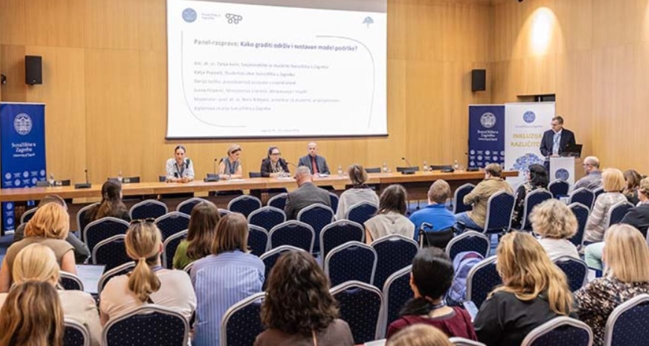 Na fotografiji je prikazan stručni skup u velikoj konferencijskoj dvorani. U prvom planu vide se sudionici koji sjede u redovima plavih stolica i prate panel-raspravu. Na pozornici za dugim stolom sjedi nekoliko panelista, dok jedan govornik stoji za govornicom s desne strane dvorane i obraća se okupljenima. Iza sudionika na pozornici nalazi se veliko projekcijsko platno s naslovom panel-rasprave „Kako graditi održiv i sustavan model podrške?“, a uz pozornicu su postavljeni promotivni roll-up plakati Sveučilišta u Zagrebu i skupa posvećenog inkluziji i različitosti. Prostor djeluje formalno i akademski, a atmosfera odražava stručni i edukativni karakter događanja.