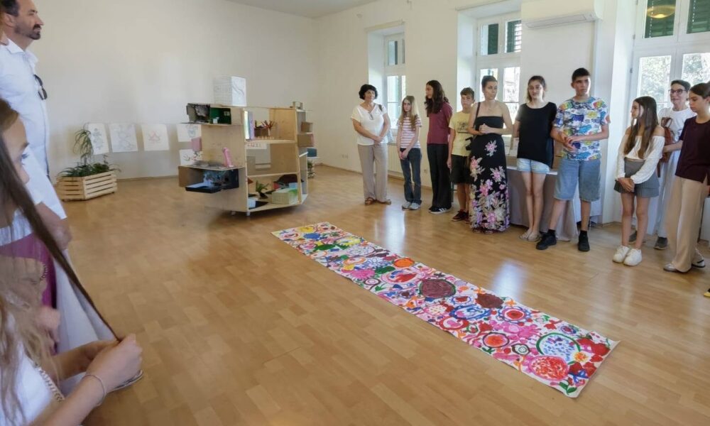 Na fotografiji je prikazan trenutak sa završne izložbe projekta „Ljepota različitosti = snaga umjetnosti“. U prostranoj i svijetloj galerijskoj dvorani okupljeni su sudionici projekta, djeca, roditelji i posjetitelji koji razgledavaju radove nastale tijekom kreativnih radionica. U središtu prostora nalazi se dugački zajednički likovni rad ispunjen živopisnim bojama, uzorcima i motivima koji odražavaju dječju maštu, kreativnost i zajedničko stvaralaštvo. Uz njega su izloženi i trodimenzionalni radovi te crteži postavljeni na zidovima i izložbenim elementima. Sudionici stoje okupljeni oko izložbe, pažljivo slušajući predstavljanje radova, dok atmosfera odiše ponosom, zajedništvom i međusobnom podrškom. Fotografija zorno prikazuje inkluzivni karakter projekta, u kojem su djeca različitih sposobnosti kroz umjetnost zajedno učila, stvarala i razvijala nova prijateljstva.