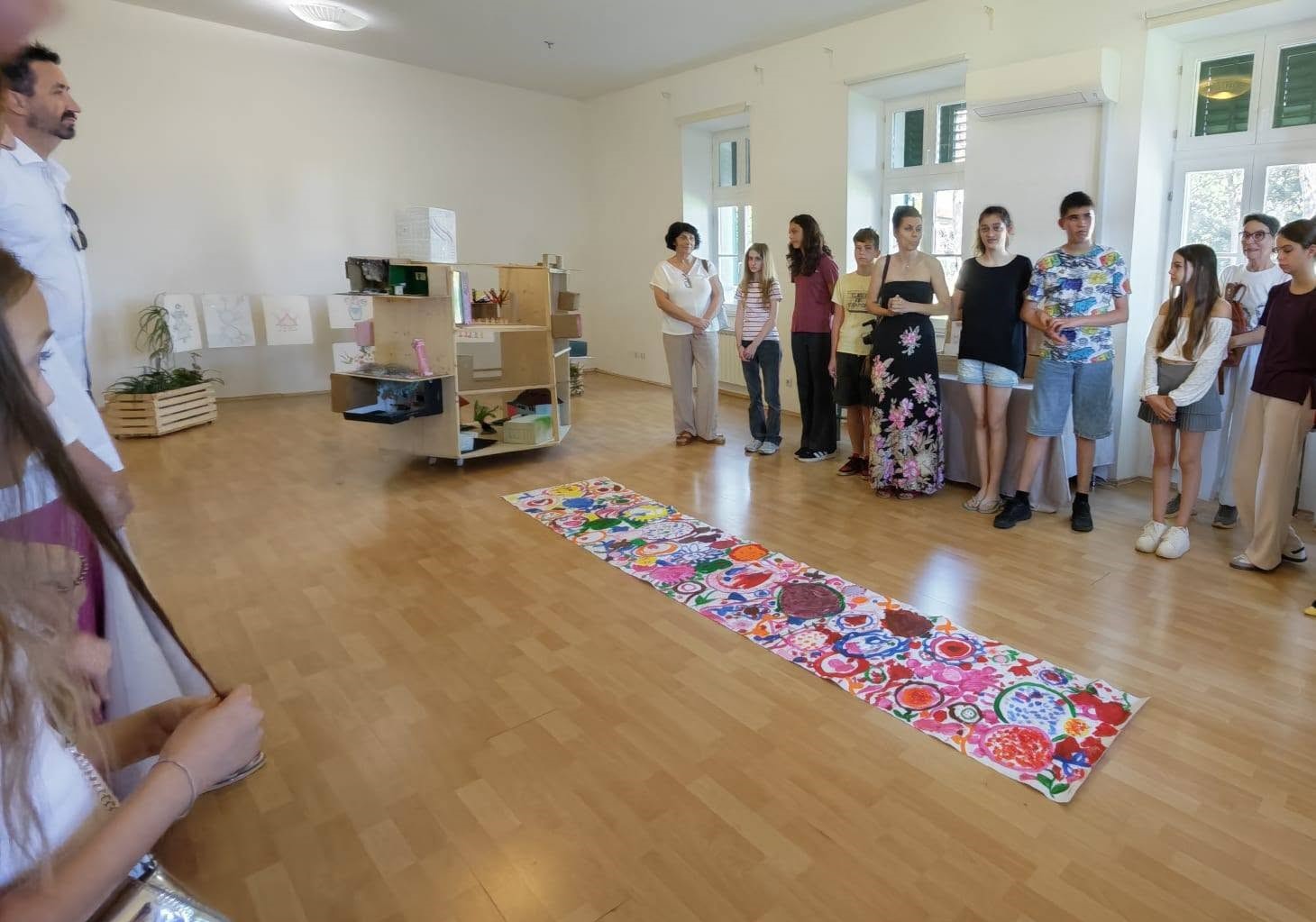 Na fotografiji je prikazan trenutak sa završne izložbe projekta „Ljepota različitosti = snaga umjetnosti“. U prostranoj i svijetloj galerijskoj dvorani okupljeni su sudionici projekta, djeca, roditelji i posjetitelji koji razgledavaju radove nastale tijekom kreativnih radionica. U središtu prostora nalazi se dugački zajednički likovni rad ispunjen živopisnim bojama, uzorcima i motivima koji odražavaju dječju maštu, kreativnost i zajedničko stvaralaštvo. Uz njega su izloženi i trodimenzionalni radovi te crteži postavljeni na zidovima i izložbenim elementima. Sudionici stoje okupljeni oko izložbe, pažljivo slušajući predstavljanje radova, dok atmosfera odiše ponosom, zajedništvom i međusobnom podrškom. Fotografija zorno prikazuje inkluzivni karakter projekta, u kojem su djeca različitih sposobnosti kroz umjetnost zajedno učila, stvarala i razvijala nova prijateljstva.