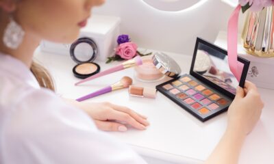 Na fotografiji se vidi osoba koja sjedi za bijelim kozmetičkim stolom ispred ogledala i pregledava otvorenu paletu sjenila za oči. Lice osobe nije u potpunosti vidljivo jer je fotografirana iz bočnog kuta i zamućenog prednjeg plana, dok je fokus stavljen na kozmetičke proizvode. Na stolu se nalaze dvije četkice za šminkanje s ružičasto-ljubičastim drškama, kompaktni puder, posudica s puderom u prahu, tekući sjajilo ili ruž za usne te otvorena paleta sjenila s raznim nijansama, od bež i smeđih do ružičastih, ljubičastih i narančastih tonova. Poklopac palete sadrži ogledalo u kojem se djelomično odražavaju proizvodi. U pozadini se vide ukrasni cvjetovi u ružičastoj i ljubičastoj boji te zlatna vaza s ružičastom vrpcom, što prostoru daje elegantan i ženstven izgled. Cijela scena odiše urednošću i svijetlim tonovima, a prirodno ili meko umjetno osvjetljenje dodatno naglašava boje kozmetike i stvara ugodnu atmosferu. Fotografija je prikladna za teme vezane uz šminkanje, kozmetiku, beauty rutinu i njegu ljepote.