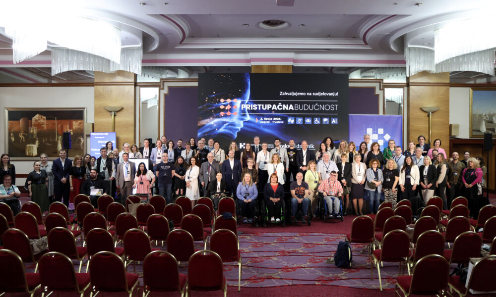Na fotografiji je prikazana velika skupina sudionika konferencije okupljena u konferencijskoj dvorani nakon završetka događaja. Ljudi stoje i sjede u nekoliko redova ispred pozornice, a među sudionicima su i osobe koje koriste invalidska kolica, što dodatno naglašava inkluzivni karakter konferencije. U pozadini dominira veliki ekran s natpisom „Pristupačna budućnost” i porukom „Zahvaljujemo na sudjelovanju!”, uz vizualne elemente povezane s tehnologijom, umjetnom inteligencijom i pristupačnošću. Na ekranu se također vide simboli za različite oblike pristupačnosti, poput invalidskih kolica, vida i sluha. Dvorana je elegantno uređena, s crvenim stolicama, velikim lusterima i dekorativnim stropom. U prvom planu nalaze se prazni redovi stolica, dok su sudionici okupljeni oko pozornice za zajedničko fotografiranje. Atmosfera djeluje profesionalno, svečano i prijateljski, a fotografija odaje dojam uspješnog i dobro posjećenog događaja.