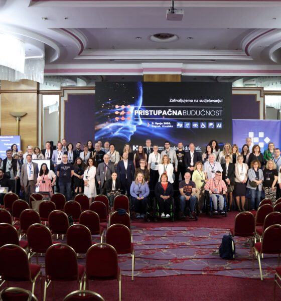 Na fotografiji je prikazana velika skupina sudionika konferencije okupljena u konferencijskoj dvorani nakon završetka događaja. Ljudi stoje i sjede u nekoliko redova ispred pozornice, a među sudionicima su i osobe koje koriste invalidska kolica, što dodatno naglašava inkluzivni karakter konferencije. U pozadini dominira veliki ekran s natpisom „Pristupačna budućnost” i porukom „Zahvaljujemo na sudjelovanju!”, uz vizualne elemente povezane s tehnologijom, umjetnom inteligencijom i pristupačnošću. Na ekranu se također vide simboli za različite oblike pristupačnosti, poput invalidskih kolica, vida i sluha. Dvorana je elegantno uređena, s crvenim stolicama, velikim lusterima i dekorativnim stropom. U prvom planu nalaze se prazni redovi stolica, dok su sudionici okupljeni oko pozornice za zajedničko fotografiranje. Atmosfera djeluje profesionalno, svečano i prijateljski, a fotografija odaje dojam uspješnog i dobro posjećenog događaja.