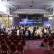 Na fotografiji je prikazana velika skupina sudionika konferencije okupljena u konferencijskoj dvorani nakon završetka događaja. Ljudi stoje i sjede u nekoliko redova ispred pozornice, a među sudionicima su i osobe koje koriste invalidska kolica, što dodatno naglašava inkluzivni karakter konferencije. U pozadini dominira veliki ekran s natpisom „Pristupačna budućnost” i porukom „Zahvaljujemo na sudjelovanju!”, uz vizualne elemente povezane s tehnologijom, umjetnom inteligencijom i pristupačnošću. Na ekranu se također vide simboli za različite oblike pristupačnosti, poput invalidskih kolica, vida i sluha. Dvorana je elegantno uređena, s crvenim stolicama, velikim lusterima i dekorativnim stropom. U prvom planu nalaze se prazni redovi stolica, dok su sudionici okupljeni oko pozornice za zajedničko fotografiranje. Atmosfera djeluje profesionalno, svečano i prijateljski, a fotografija odaje dojam uspješnog i dobro posjećenog događaja.
