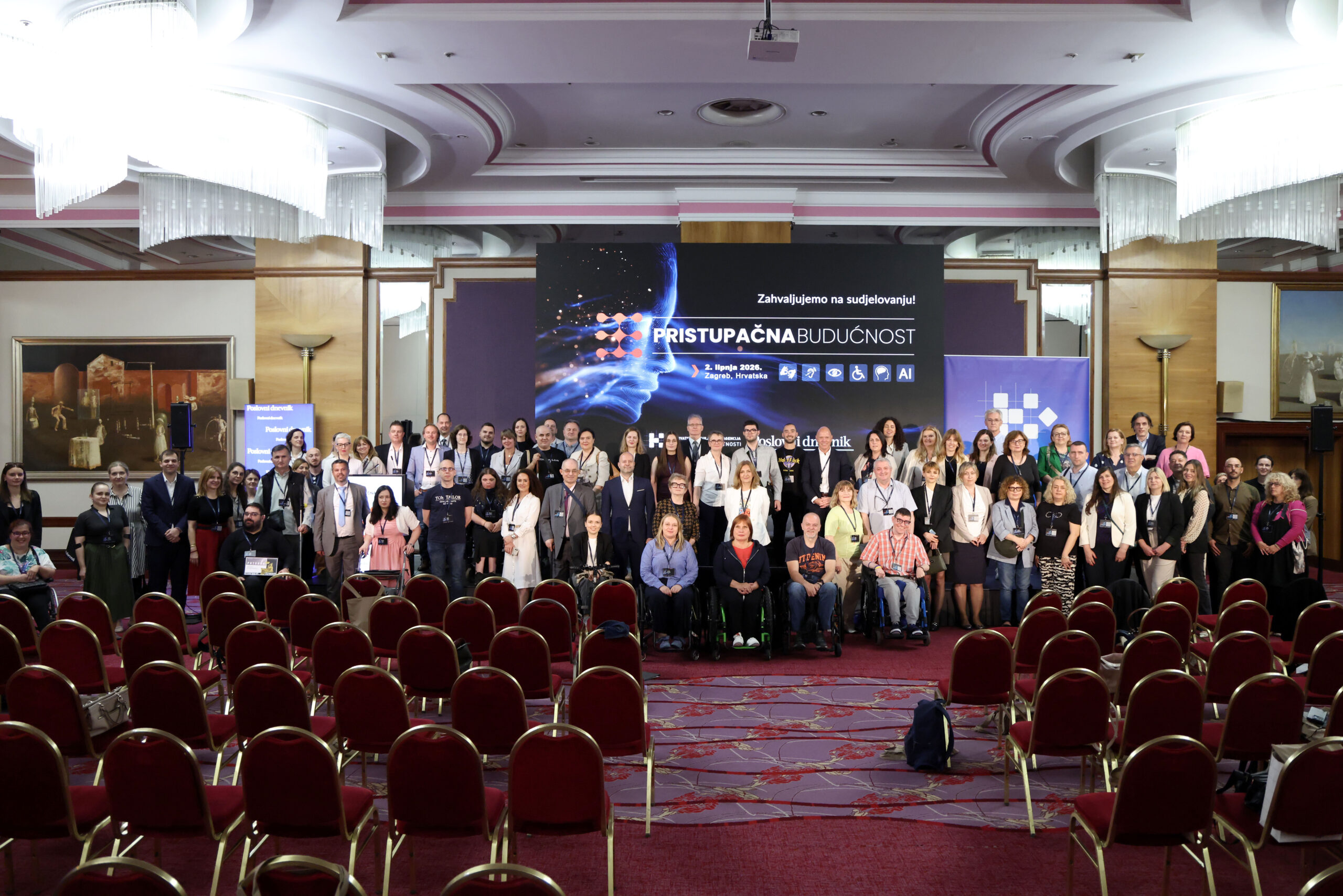 Na fotografiji je prikazana velika skupina sudionika konferencije okupljena u konferencijskoj dvorani nakon završetka događaja. Ljudi stoje i sjede u nekoliko redova ispred pozornice, a među sudionicima su i osobe koje koriste invalidska kolica, što dodatno naglašava inkluzivni karakter konferencije. U pozadini dominira veliki ekran s natpisom „Pristupačna budućnost” i porukom „Zahvaljujemo na sudjelovanju!”, uz vizualne elemente povezane s tehnologijom, umjetnom inteligencijom i pristupačnošću. Na ekranu se također vide simboli za različite oblike pristupačnosti, poput invalidskih kolica, vida i sluha. Dvorana je elegantno uređena, s crvenim stolicama, velikim lusterima i dekorativnim stropom. U prvom planu nalaze se prazni redovi stolica, dok su sudionici okupljeni oko pozornice za zajedničko fotografiranje. Atmosfera djeluje profesionalno, svečano i prijateljski, a fotografija odaje dojam uspješnog i dobro posjećenog događaja.