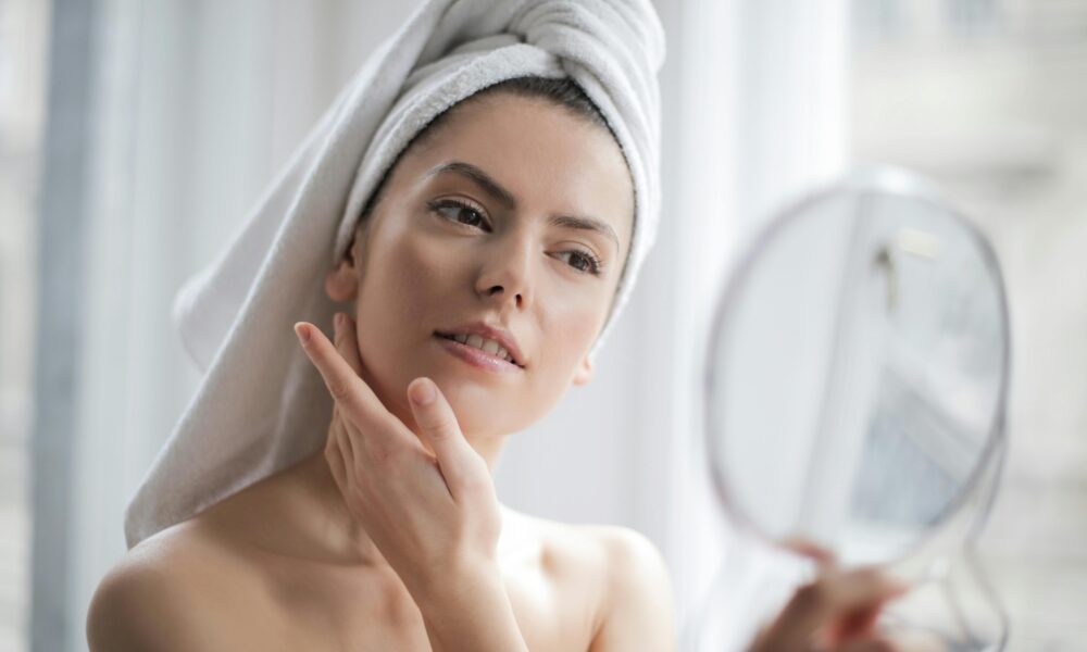 Na fotografiji je prikazana mlada žena koja stoji ispred ogledala tijekom svoje rutine njege kože. Kosa joj je omotana u bijeli ručnik, što sugerira da je fotografija snimljena neposredno nakon tuširanja ili pranja kose. Nježno dodiruje lice i vrat, promatrajući svoj odraz u okruglom ogledalu, dok na licu ima prirodan, svjež izgled bez naglašene šminke. Izraz lica djeluje opušteno i zadovoljno, stvarajući dojam samopouzdanja i brige o vlastitoj koži. Pozadina je svijetla i minimalistički uređena, s puno prirodnog svjetla koje dodatno naglašava čistoću i svježinu prizora. Cjelokupna fotografija odiše osjećajem njege, opuštanja i wellness atmosfere te se često koristi za ilustriranje tema poput svakodnevne beauty rutine, njege kože, hidratacije, prirodne ljepote i zdravog načina života.