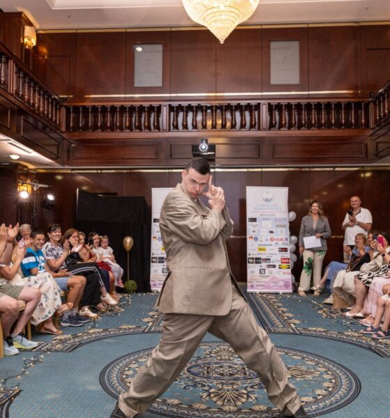 Na fotografiji je prikazana modna revija održana u elegantnoj hotelskoj dvorani, gdje središnjim prostorom između publike samouvjereno prolazi mladi muškarac odjeven u široko krojeno svijetlosmeđe odijelo. U dinamičnoj pozi privlači pažnju okupljenih gledatelja koji ga nagrađuju pljeskom i osmijesima, stvarajući atmosferu podrške, ponosa i zajedništva. Publika sjedi s obje strane improvizirane modne piste, dok raskošni interijer s tamnim drvenim detaljima, galerijama i velikim kristalnim lusterom dodatno naglašava svečanost događaja. U pozadini su vidljivi promotivni panoi organizatora i partnera, a cijeli prostor odiše toplinom i pozitivnom energijom. Fotografija uspješno prenosi duh inkluzije, samopouzdanja i osobnog izražavanja te prikazuje trenutak u kojem sudionik revije s ponosom pokazuje svoju osobnost pred publikom koja ga srdačno podržava.