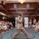 Na fotografiji je prikazana modna revija održana u elegantnoj hotelskoj dvorani, gdje središnjim prostorom između publike samouvjereno prolazi mladi muškarac odjeven u široko krojeno svijetlosmeđe odijelo. U dinamičnoj pozi privlači pažnju okupljenih gledatelja koji ga nagrađuju pljeskom i osmijesima, stvarajući atmosferu podrške, ponosa i zajedništva. Publika sjedi s obje strane improvizirane modne piste, dok raskošni interijer s tamnim drvenim detaljima, galerijama i velikim kristalnim lusterom dodatno naglašava svečanost događaja. U pozadini su vidljivi promotivni panoi organizatora i partnera, a cijeli prostor odiše toplinom i pozitivnom energijom. Fotografija uspješno prenosi duh inkluzije, samopouzdanja i osobnog izražavanja te prikazuje trenutak u kojem sudionik revije s ponosom pokazuje svoju osobnost pred publikom koja ga srdačno podržava.