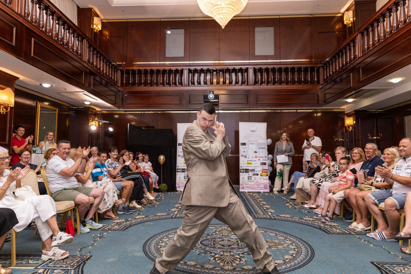 Na fotografiji je prikazana modna revija održana u elegantnoj hotelskoj dvorani, gdje središnjim prostorom između publike samouvjereno prolazi mladi muškarac odjeven u široko krojeno svijetlosmeđe odijelo. U dinamičnoj pozi privlači pažnju okupljenih gledatelja koji ga nagrađuju pljeskom i osmijesima, stvarajući atmosferu podrške, ponosa i zajedništva. Publika sjedi s obje strane improvizirane modne piste, dok raskošni interijer s tamnim drvenim detaljima, galerijama i velikim kristalnim lusterom dodatno naglašava svečanost događaja. U pozadini su vidljivi promotivni panoi organizatora i partnera, a cijeli prostor odiše toplinom i pozitivnom energijom. Fotografija uspješno prenosi duh inkluzije, samopouzdanja i osobnog izražavanja te prikazuje trenutak u kojem sudionik revije s ponosom pokazuje svoju osobnost pred publikom koja ga srdačno podržava.