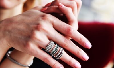 Na slici se vidi ženska ruka s nekoliko elegantnih prstenova složenih jedan do drugoga na prstu, što predstavlja popularni trend ring stackinga. Prstenovi su srebrne boje i ukrašeni sjajnim kamenčićima različitih boja, zbog čega izgledaju luksuzno i moderno. Nokti su uredno nalakirani svijetloružičastom bojom, a na zapešću se nalazi jednostavna narukvica. U pozadini se nazire crvena fotelja i dio osobe, dok je fokus fotografije stavljen na nakit i ruke. Cijela slika ostavlja elegantan i profinjen dojam te naglašava modu i stil nošenja više prstena odjednom.