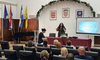 Na fotografiji je prikazan trenutak s Dana otvorenih vrata Udruge Vretenac u dvorani Hrvatskog doma u Slatini. U prvom planu nalaze se okupljeni posjetitelji koji prate predavanje, dok u središtu prostorije predavačica stoji ispred projekcijskog platna i obraća se sudionicima. Na platnu je prikazana naslovna prezentacija događaja „100 lica inkluzije: Svi za jednog, zajednica za sve!“. Prostor je svečano uređen, s cvjetnim vijencem iznad pozornice te istaknutim zastavama Republike Hrvatske, Europske unije, Virovitičko-podravske županije i Grada Slatine. Na zidu iza pozornice nalaze se grbovi Grada Slatine, Republike Hrvatske i Virovitičko-podravske županije. Fotografija dočarava radnu i ugodnu atmosferu događaja, uz aktivno sudjelovanje okupljenih građana i predstavnika lokalne zajednice.