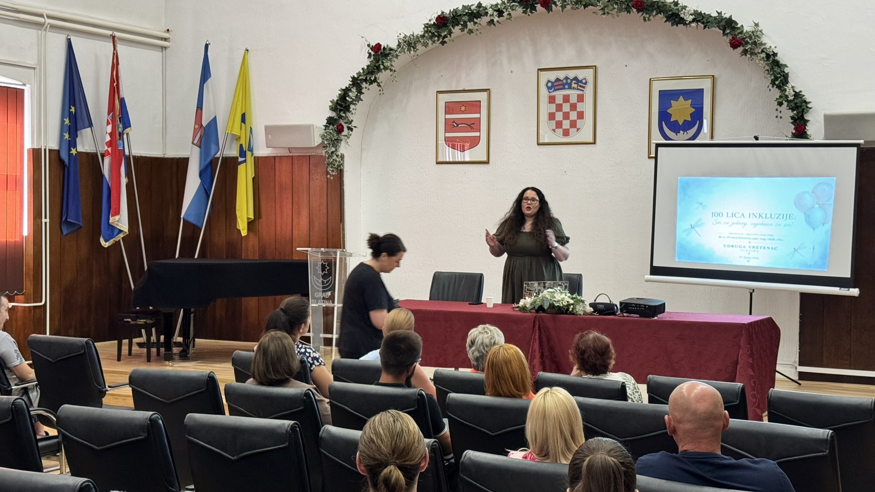 Na fotografiji je prikazan trenutak s Dana otvorenih vrata Udruge Vretenac u dvorani Hrvatskog doma u Slatini. U prvom planu nalaze se okupljeni posjetitelji koji prate predavanje, dok u središtu prostorije predavačica stoji ispred projekcijskog platna i obraća se sudionicima. Na platnu je prikazana naslovna prezentacija događaja „100 lica inkluzije: Svi za jednog, zajednica za sve!“. Prostor je svečano uređen, s cvjetnim vijencem iznad pozornice te istaknutim zastavama Republike Hrvatske, Europske unije, Virovitičko-podravske županije i Grada Slatine. Na zidu iza pozornice nalaze se grbovi Grada Slatine, Republike Hrvatske i Virovitičko-podravske županije. Fotografija dočarava radnu i ugodnu atmosferu događaja, uz aktivno sudjelovanje okupljenih građana i predstavnika lokalne zajednice.