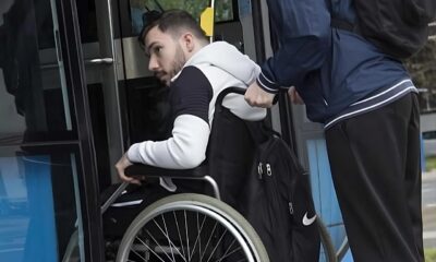 Na fotografiji se vidi osoba u invalidskim kolicima kojoj druga osoba pomaže pri ulasku u niskopodni autobus. Osoba u kolicima nalazi se na ulazu u vozilo i jednom rukom pridržava okvir vrata, dok je iza nje osoba koja gura kolica i pomaže pri ukrcaju. Fotografija naglašava važnost pristupačnog javnog prijevoza i uloge osobne asistencije u omogućavanju samostalnijeg kretanja osoba s invaliditetom. Prikazuje svakodnevnu situaciju u kojoj prilagođena vozila i podrška druge osobe omogućuju lakše korištenje javnog prijevoza.