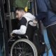 Na fotografiji se vidi osoba u invalidskim kolicima kojoj druga osoba pomaže pri ulasku u niskopodni autobus. Osoba u kolicima nalazi se na ulazu u vozilo i jednom rukom pridržava okvir vrata, dok je iza nje osoba koja gura kolica i pomaže pri ukrcaju. Fotografija naglašava važnost pristupačnog javnog prijevoza i uloge osobne asistencije u omogućavanju samostalnijeg kretanja osoba s invaliditetom. Prikazuje svakodnevnu situaciju u kojoj prilagođena vozila i podrška druge osobe omogućuju lakše korištenje javnog prijevoza.