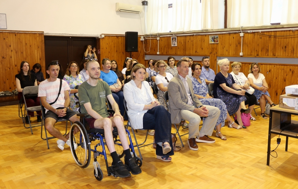 Na fotografiji je prikazan skup povodom obilježavanja obljetnice Centra za pružanje usluga u zajednici Varaždin. U prvom planu sjede uzvanici i korisnici Centra, među kojima je i osoba u invalidskim kolicima, što dodatno naglašava inkluzivni karakter događanja. Sudionici pažljivo prate program koji se odvija u dvorani. Prostorija je ispunjena brojnim posjetiteljima, a u pozadini se mogu vidjeti redovi sjedećih mjesta i osoba koja fotografira događaj. Topla i svečana atmosfera odražava zajedništvo, podršku i važnost rada Centra za lokalnu zajednicu.