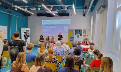 Na fotografiji je prikazan skup sudionika na početnoj konferenciji projekta „Zajednica jednakih mogućnosti – socijalne usluge dostupne svima“. U moderno uređenoj dvorani okupljeni sjede i prate izlaganje koje se održava ispred projekcijskog platna na kojem je istaknut naziv projekta te logotipi Društva za istraživanje i potporu (DIP) i Primorsko-goranske županije. U prvom planu vide se sudionici konferencije koji sjede na žutim i zelenim stolicama, dok nekoliko osoba stoji sa strane. Na pozornici se nalazi govornica koja se obraća okupljenima, a iza nje sjede još tri osobe, vjerojatno članovi projektnog tima ili panelisti. S lijeve strane postavljena je kamera koja snima događanje, dok se s desne strane nalaze promotivni roll-up panoi. Fotografija prikazuje radnu i svečanu atmosferu konferencije te naglašava suradnju institucija i organizacija uključenih u razvoj dostupnijih socijalnih usluga za osobe s invaliditetom i njihove obitelji u Primorsko-goranskoj županiji.