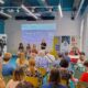 Na fotografiji je prikazan skup sudionika na početnoj konferenciji projekta „Zajednica jednakih mogućnosti – socijalne usluge dostupne svima“. U moderno uređenoj dvorani okupljeni sjede i prate izlaganje koje se održava ispred projekcijskog platna na kojem je istaknut naziv projekta te logotipi Društva za istraživanje i potporu (DIP) i Primorsko-goranske županije. U prvom planu vide se sudionici konferencije koji sjede na žutim i zelenim stolicama, dok nekoliko osoba stoji sa strane. Na pozornici se nalazi govornica koja se obraća okupljenima, a iza nje sjede još tri osobe, vjerojatno članovi projektnog tima ili panelisti. S lijeve strane postavljena je kamera koja snima događanje, dok se s desne strane nalaze promotivni roll-up panoi. Fotografija prikazuje radnu i svečanu atmosferu konferencije te naglašava suradnju institucija i organizacija uključenih u razvoj dostupnijih socijalnih usluga za osobe s invaliditetom i njihove obitelji u Primorsko-goranskoj županiji.