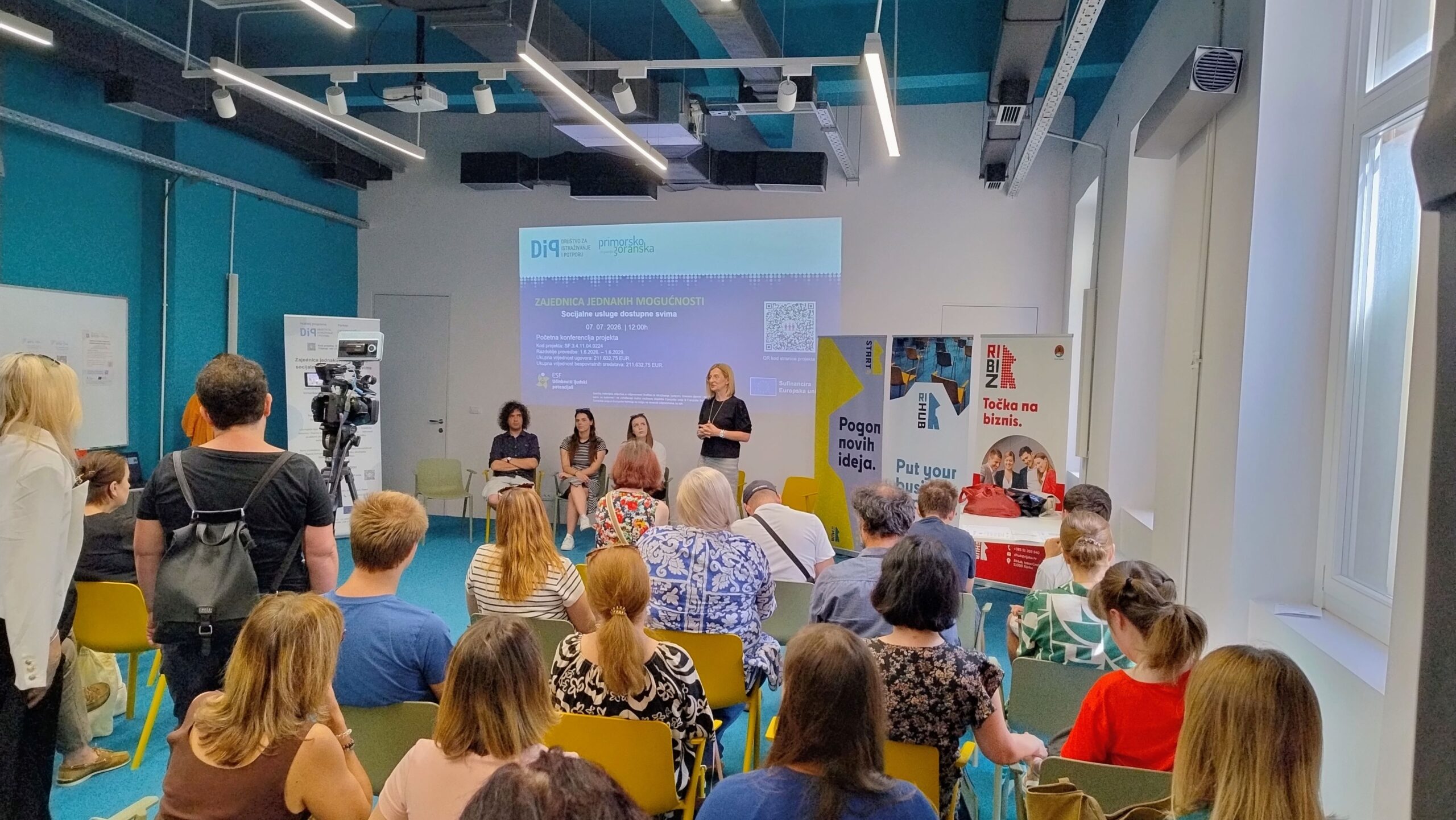 Na fotografiji je prikazan skup sudionika na početnoj konferenciji projekta „Zajednica jednakih mogućnosti – socijalne usluge dostupne svima“. U moderno uređenoj dvorani okupljeni sjede i prate izlaganje koje se održava ispred projekcijskog platna na kojem je istaknut naziv projekta te logotipi Društva za istraživanje i potporu (DIP) i Primorsko-goranske županije. U prvom planu vide se sudionici konferencije koji sjede na žutim i zelenim stolicama, dok nekoliko osoba stoji sa strane. Na pozornici se nalazi govornica koja se obraća okupljenima, a iza nje sjede još tri osobe, vjerojatno članovi projektnog tima ili panelisti. S lijeve strane postavljena je kamera koja snima događanje, dok se s desne strane nalaze promotivni roll-up panoi. Fotografija prikazuje radnu i svečanu atmosferu konferencije te naglašava suradnju institucija i organizacija uključenih u razvoj dostupnijih socijalnih usluga za osobe s invaliditetom i njihove obitelji u Primorsko-goranskoj županiji.