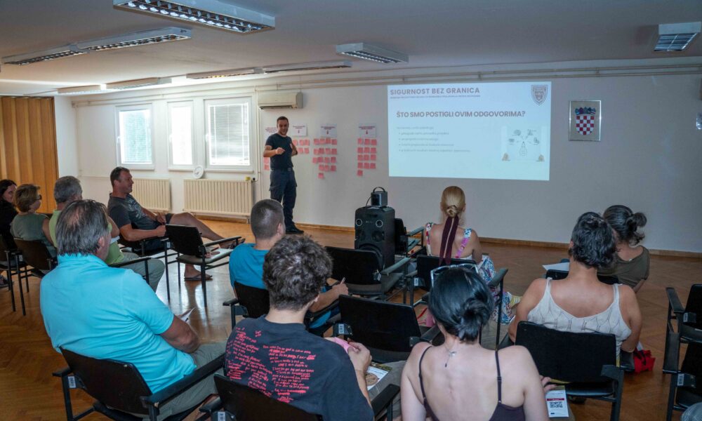 Na fotografiji je prikazan radionički sastanak projekta „Sigurnost bez granica“ u dvorani za predavanja. U prvom planu vide se sudionici koji sjede u redovima i prate prezentaciju, dok predavač stoji ispred projekcijskog platna i predstavlja rezultate i aktivnosti projekta. Na zidu je projicirana prezentacija s naslovom „Sigurnost bez granica“, a uz platno su postavljeni plakati s bilješkama sudionika, što upućuje na interaktivni dio radionice i prikupljanje prijedloga građana. Fotografija prikazuje aktivno sudjelovanje lokalne zajednice u raspravi o prevenciji požara i održivim alternativama spaljivanju biljnog otpada.
