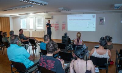 Na fotografiji je prikazan radionički sastanak projekta „Sigurnost bez granica“ u dvorani za predavanja. U prvom planu vide se sudionici koji sjede u redovima i prate prezentaciju, dok predavač stoji ispred projekcijskog platna i predstavlja rezultate i aktivnosti projekta. Na zidu je projicirana prezentacija s naslovom „Sigurnost bez granica“, a uz platno su postavljeni plakati s bilješkama sudionika, što upućuje na interaktivni dio radionice i prikupljanje prijedloga građana. Fotografija prikazuje aktivno sudjelovanje lokalne zajednice u raspravi o prevenciji požara i održivim alternativama spaljivanju biljnog otpada.