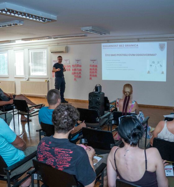 Na fotografiji je prikazan radionički sastanak projekta „Sigurnost bez granica“ u dvorani za predavanja. U prvom planu vide se sudionici koji sjede u redovima i prate prezentaciju, dok predavač stoji ispred projekcijskog platna i predstavlja rezultate i aktivnosti projekta. Na zidu je projicirana prezentacija s naslovom „Sigurnost bez granica“, a uz platno su postavljeni plakati s bilješkama sudionika, što upućuje na interaktivni dio radionice i prikupljanje prijedloga građana. Fotografija prikazuje aktivno sudjelovanje lokalne zajednice u raspravi o prevenciji požara i održivim alternativama spaljivanju biljnog otpada.