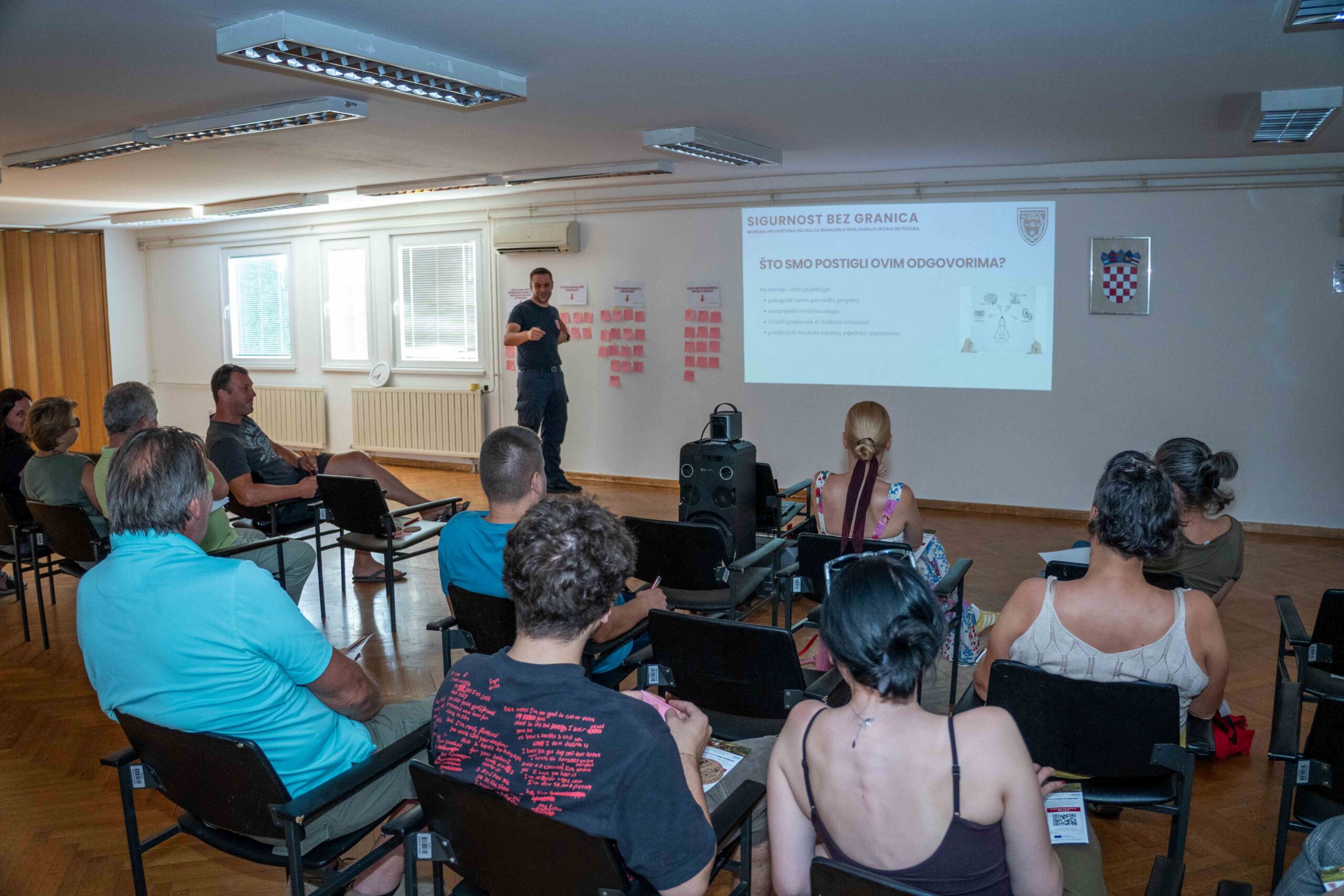 Na fotografiji je prikazan radionički sastanak projekta „Sigurnost bez granica“ u dvorani za predavanja. U prvom planu vide se sudionici koji sjede u redovima i prate prezentaciju, dok predavač stoji ispred projekcijskog platna i predstavlja rezultate i aktivnosti projekta. Na zidu je projicirana prezentacija s naslovom „Sigurnost bez granica“, a uz platno su postavljeni plakati s bilješkama sudionika, što upućuje na interaktivni dio radionice i prikupljanje prijedloga građana. Fotografija prikazuje aktivno sudjelovanje lokalne zajednice u raspravi o prevenciji požara i održivim alternativama spaljivanju biljnog otpada.