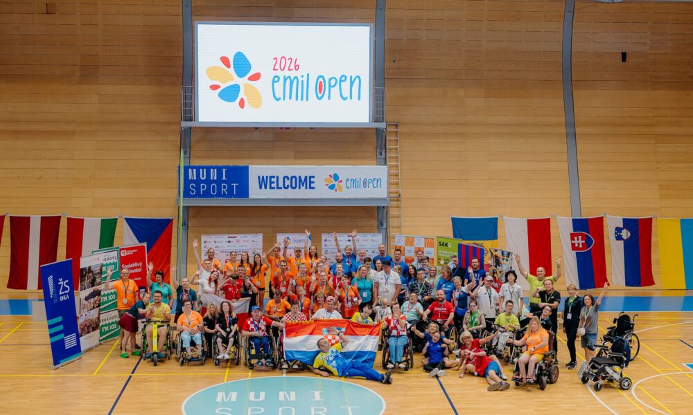 Na fotografiji je zabilježen zajednički trenutak sudionika međunarodnog sportskog događaja Emil Open 2026, održanog u sportskoj dvorani. U središtu fotografije okupljeni su sportaši, treneri, volonteri i članovi organizacije koji poziraju nakon natjecanja. Mnogi sudionici podižu ruke i osmjehuju se, čime se ističe vesela i svečana atmosfera. U prvom planu nalazi se skupina hrvatskih sudionika koji ponosno drže hrvatsku zastavu, a oko njih su sportaši u invalidskim kolicima i ostali članovi reprezentacija. Većina sudionika nosi službene majice različitih boja, akreditacije i medalje oko vrata, što upućuje na završetak natjecanja ili ceremoniju proglašenja. U pozadini se nalazi veliki digitalni ekran s logotipom Emil Open 2026, dok ispod njega stoji natpis dobrodošlice. Dvorana je ukrašena zastavama više europskih zemalja, što dodatno naglašava međunarodni karakter događaja. Fotografija odiše zajedništvom, sportskim duhom i slavljem uspjeha te prikazuje inkluzivnost i prijateljsku atmosferu koja je obilježila natjecanje.