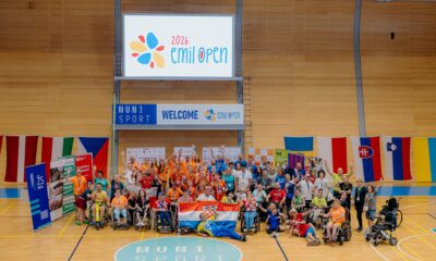 Na fotografiji je zabilježen zajednički trenutak sudionika međunarodnog sportskog događaja Emil Open 2026, održanog u sportskoj dvorani. U središtu fotografije okupljeni su sportaši, treneri, volonteri i članovi organizacije koji poziraju nakon natjecanja. Mnogi sudionici podižu ruke i osmjehuju se, čime se ističe vesela i svečana atmosfera. U prvom planu nalazi se skupina hrvatskih sudionika koji ponosno drže hrvatsku zastavu, a oko njih su sportaši u invalidskim kolicima i ostali članovi reprezentacija. Većina sudionika nosi službene majice različitih boja, akreditacije i medalje oko vrata, što upućuje na završetak natjecanja ili ceremoniju proglašenja. U pozadini se nalazi veliki digitalni ekran s logotipom Emil Open 2026, dok ispod njega stoji natpis dobrodošlice. Dvorana je ukrašena zastavama više europskih zemalja, što dodatno naglašava međunarodni karakter događaja. Fotografija odiše zajedništvom, sportskim duhom i slavljem uspjeha te prikazuje inkluzivnost i prijateljsku atmosferu koja je obilježila natjecanje.