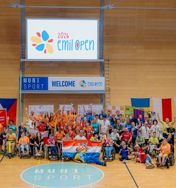 Na fotografiji je zabilježen zajednički trenutak sudionika međunarodnog sportskog događaja Emil Open 2026, održanog u sportskoj dvorani. U središtu fotografije okupljeni su sportaši, treneri, volonteri i članovi organizacije koji poziraju nakon natjecanja. Mnogi sudionici podižu ruke i osmjehuju se, čime se ističe vesela i svečana atmosfera. U prvom planu nalazi se skupina hrvatskih sudionika koji ponosno drže hrvatsku zastavu, a oko njih su sportaši u invalidskim kolicima i ostali članovi reprezentacija. Većina sudionika nosi službene majice različitih boja, akreditacije i medalje oko vrata, što upućuje na završetak natjecanja ili ceremoniju proglašenja. U pozadini se nalazi veliki digitalni ekran s logotipom Emil Open 2026, dok ispod njega stoji natpis dobrodošlice. Dvorana je ukrašena zastavama više europskih zemalja, što dodatno naglašava međunarodni karakter događaja. Fotografija odiše zajedništvom, sportskim duhom i slavljem uspjeha te prikazuje inkluzivnost i prijateljsku atmosferu koja je obilježila natjecanje.