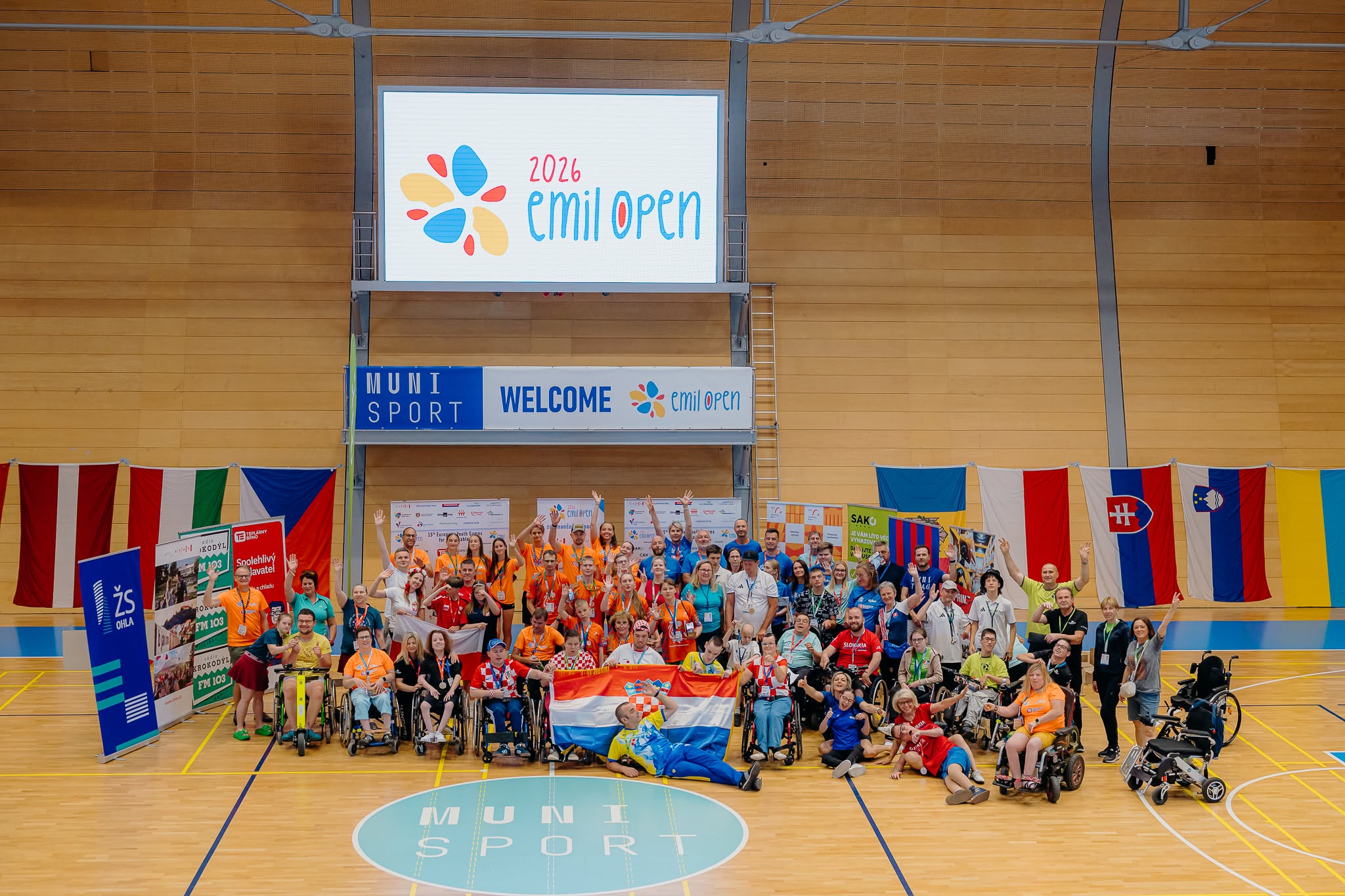 Na fotografiji je zabilježen zajednički trenutak sudionika međunarodnog sportskog događaja Emil Open 2026, održanog u sportskoj dvorani. U središtu fotografije okupljeni su sportaši, treneri, volonteri i članovi organizacije koji poziraju nakon natjecanja. Mnogi sudionici podižu ruke i osmjehuju se, čime se ističe vesela i svečana atmosfera. U prvom planu nalazi se skupina hrvatskih sudionika koji ponosno drže hrvatsku zastavu, a oko njih su sportaši u invalidskim kolicima i ostali članovi reprezentacija. Većina sudionika nosi službene majice različitih boja, akreditacije i medalje oko vrata, što upućuje na završetak natjecanja ili ceremoniju proglašenja. U pozadini se nalazi veliki digitalni ekran s logotipom Emil Open 2026, dok ispod njega stoji natpis dobrodošlice. Dvorana je ukrašena zastavama više europskih zemalja, što dodatno naglašava međunarodni karakter događaja. Fotografija odiše zajedništvom, sportskim duhom i slavljem uspjeha te prikazuje inkluzivnost i prijateljsku atmosferu koja je obilježila natjecanje.