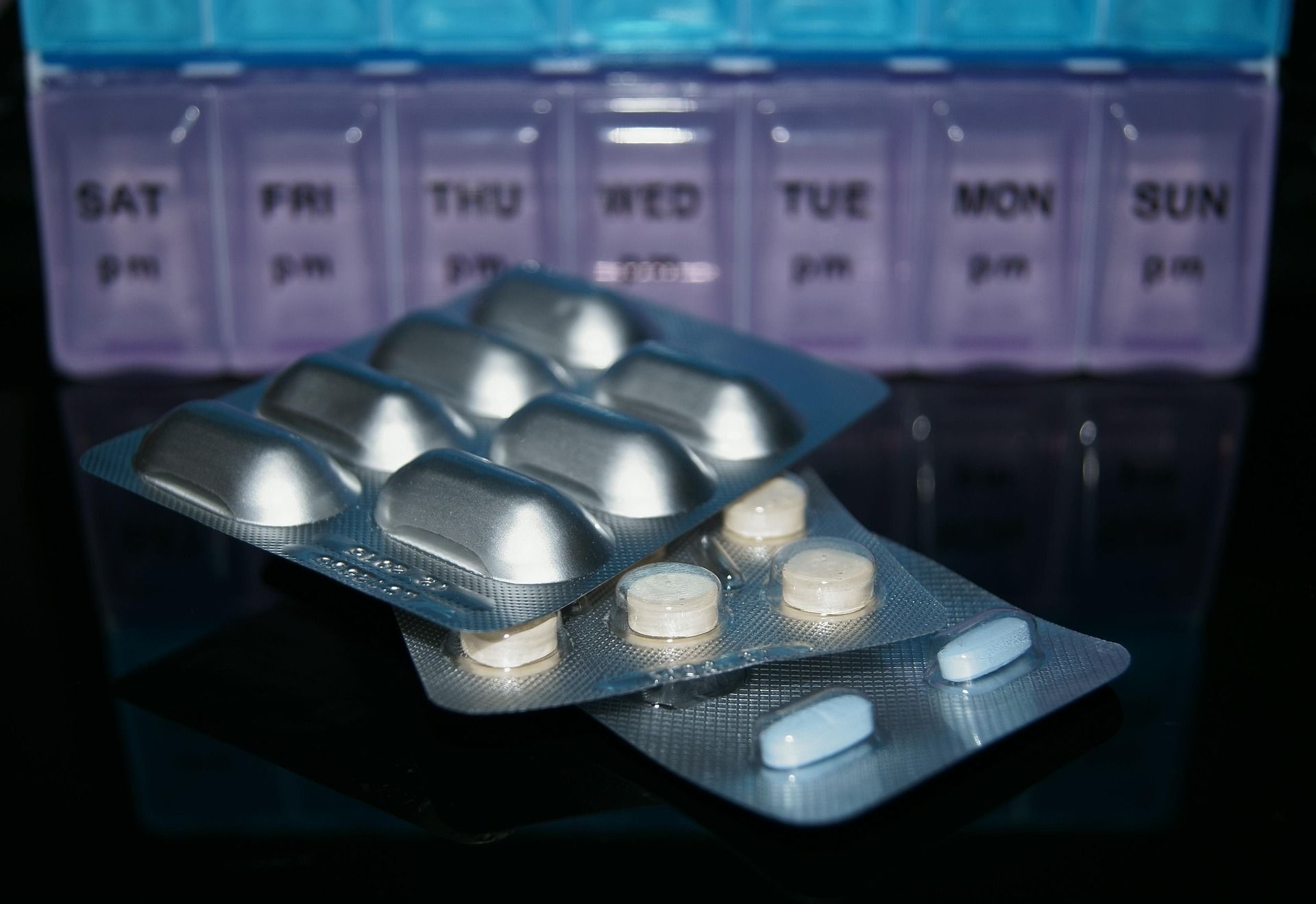 Plastična tjedna kutija za organizaciju lijekova nalazi se u pozadini, dok su u prvom planu blister pakiranja s tabletama različitih oblika. Fotografija simbolizira redovito uzimanje terapije i organizaciju lijekova.