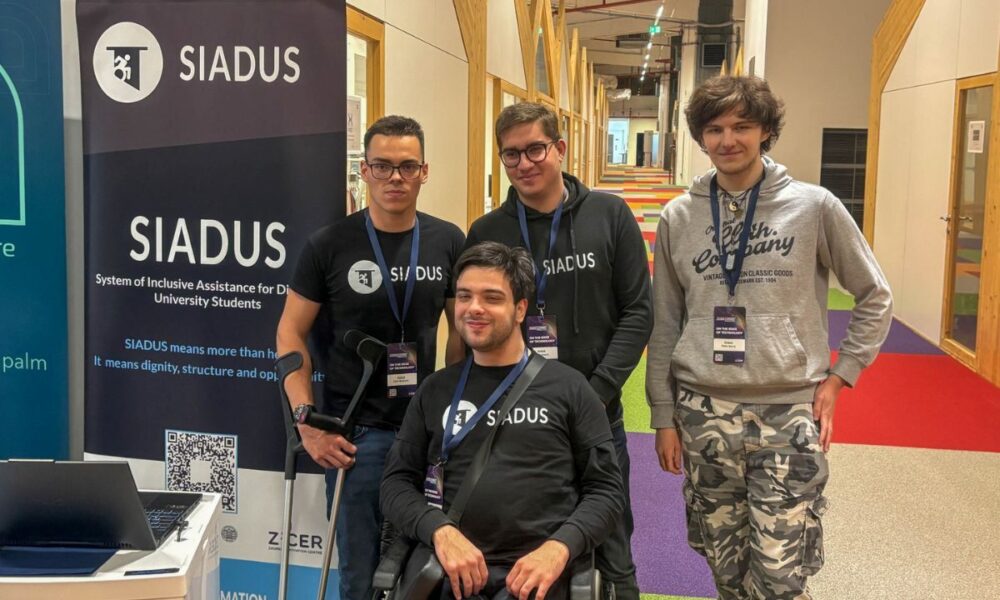 Na fotografiji je četvero članova tima SIADUS na predstavljanju projekta u modernom unutarnjem prostoru, vjerojatno tijekom startup ili inovacijskog događanja. U prvom planu nalazi se član tima u invalidskim kolicima, odjeven u crnu SIADUS majicu, s akreditacijom oko vrata. Iza njega stoje trojica kolega, također s akreditacijama, od kojih dvojica nose crne majice ili hoodice s logotipom SIADUS-a. S lijeve strane fotografije nalazi se promotivni roll-up s logotipom i nazivom SIADUS (System of Inclusive Assistance for Disabled University Students) te QR kodom za više informacija o projektu. U donjem dijelu bannera vidljiv je i logotip ZICER-a. U pozadini se pruža suvremeni hodnik s drvenim konstrukcijama, staklenim uredima i šarenim tepihom, što upućuje na prostor namijenjen inovacijama, poduzetništvu ili konferencijama. Fotografija prikazuje tim koji zajednički predstavlja SIADUS – digitalnu platformu za organizaciju podrške studentima s invaliditetom tijekom studiranja.