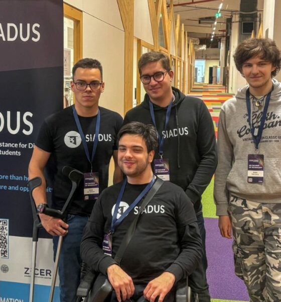 Na fotografiji je četvero članova tima SIADUS na predstavljanju projekta u modernom unutarnjem prostoru, vjerojatno tijekom startup ili inovacijskog događanja. U prvom planu nalazi se član tima u invalidskim kolicima, odjeven u crnu SIADUS majicu, s akreditacijom oko vrata. Iza njega stoje trojica kolega, također s akreditacijama, od kojih dvojica nose crne majice ili hoodice s logotipom SIADUS-a. S lijeve strane fotografije nalazi se promotivni roll-up s logotipom i nazivom SIADUS (System of Inclusive Assistance for Disabled University Students) te QR kodom za više informacija o projektu. U donjem dijelu bannera vidljiv je i logotip ZICER-a. U pozadini se pruža suvremeni hodnik s drvenim konstrukcijama, staklenim uredima i šarenim tepihom, što upućuje na prostor namijenjen inovacijama, poduzetništvu ili konferencijama. Fotografija prikazuje tim koji zajednički predstavlja SIADUS – digitalnu platformu za organizaciju podrške studentima s invaliditetom tijekom studiranja.