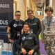 Na fotografiji je četvero članova tima SIADUS na predstavljanju projekta u modernom unutarnjem prostoru, vjerojatno tijekom startup ili inovacijskog događanja. U prvom planu nalazi se član tima u invalidskim kolicima, odjeven u crnu SIADUS majicu, s akreditacijom oko vrata. Iza njega stoje trojica kolega, također s akreditacijama, od kojih dvojica nose crne majice ili hoodice s logotipom SIADUS-a. S lijeve strane fotografije nalazi se promotivni roll-up s logotipom i nazivom SIADUS (System of Inclusive Assistance for Disabled University Students) te QR kodom za više informacija o projektu. U donjem dijelu bannera vidljiv je i logotip ZICER-a. U pozadini se pruža suvremeni hodnik s drvenim konstrukcijama, staklenim uredima i šarenim tepihom, što upućuje na prostor namijenjen inovacijama, poduzetništvu ili konferencijama. Fotografija prikazuje tim koji zajednički predstavlja SIADUS – digitalnu platformu za organizaciju podrške studentima s invaliditetom tijekom studiranja.