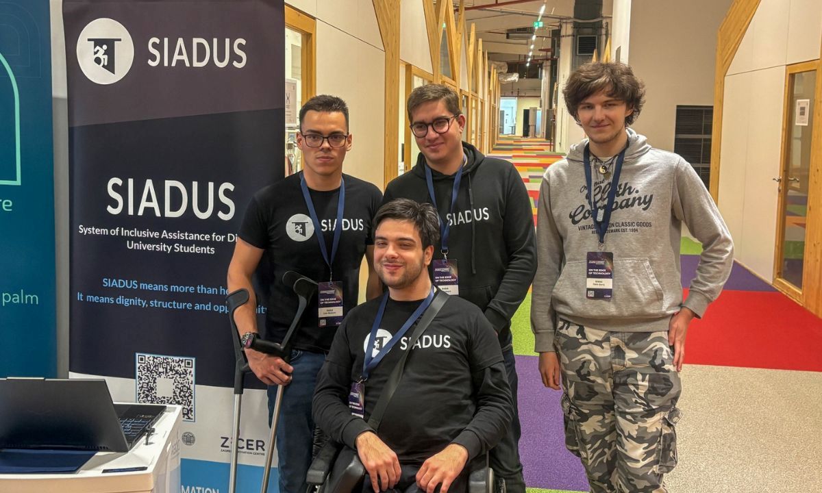 Na fotografiji je četvero članova tima SIADUS na predstavljanju projekta u modernom unutarnjem prostoru, vjerojatno tijekom startup ili inovacijskog događanja. U prvom planu nalazi se član tima u invalidskim kolicima, odjeven u crnu SIADUS majicu, s akreditacijom oko vrata. Iza njega stoje trojica kolega, također s akreditacijama, od kojih dvojica nose crne majice ili hoodice s logotipom SIADUS-a. S lijeve strane fotografije nalazi se promotivni roll-up s logotipom i nazivom SIADUS (System of Inclusive Assistance for Disabled University Students) te QR kodom za više informacija o projektu. U donjem dijelu bannera vidljiv je i logotip ZICER-a. U pozadini se pruža suvremeni hodnik s drvenim konstrukcijama, staklenim uredima i šarenim tepihom, što upućuje na prostor namijenjen inovacijama, poduzetništvu ili konferencijama. Fotografija prikazuje tim koji zajednički predstavlja SIADUS – digitalnu platformu za organizaciju podrške studentima s invaliditetom tijekom studiranja.