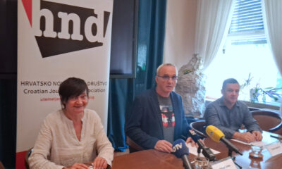 Tri osobe sjede za stolom tijekom konferencije za medije u prostorijama Hrvatskog novinarskog društva. Ispred govornika nalaze se mikrofoni više medijskih kuća, a u pozadini je istaknut banner HND-a. Sudionici odgovaraju na pitanja novinara za okruglim konferencijskim stolom.