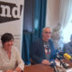 Tri osobe sjede za stolom tijekom konferencije za medije u prostorijama Hrvatskog novinarskog društva. Ispred govornika nalaze se mikrofoni više medijskih kuća, a u pozadini je istaknut banner HND-a. Sudionici odgovaraju na pitanja novinara za okruglim konferencijskim stolom.