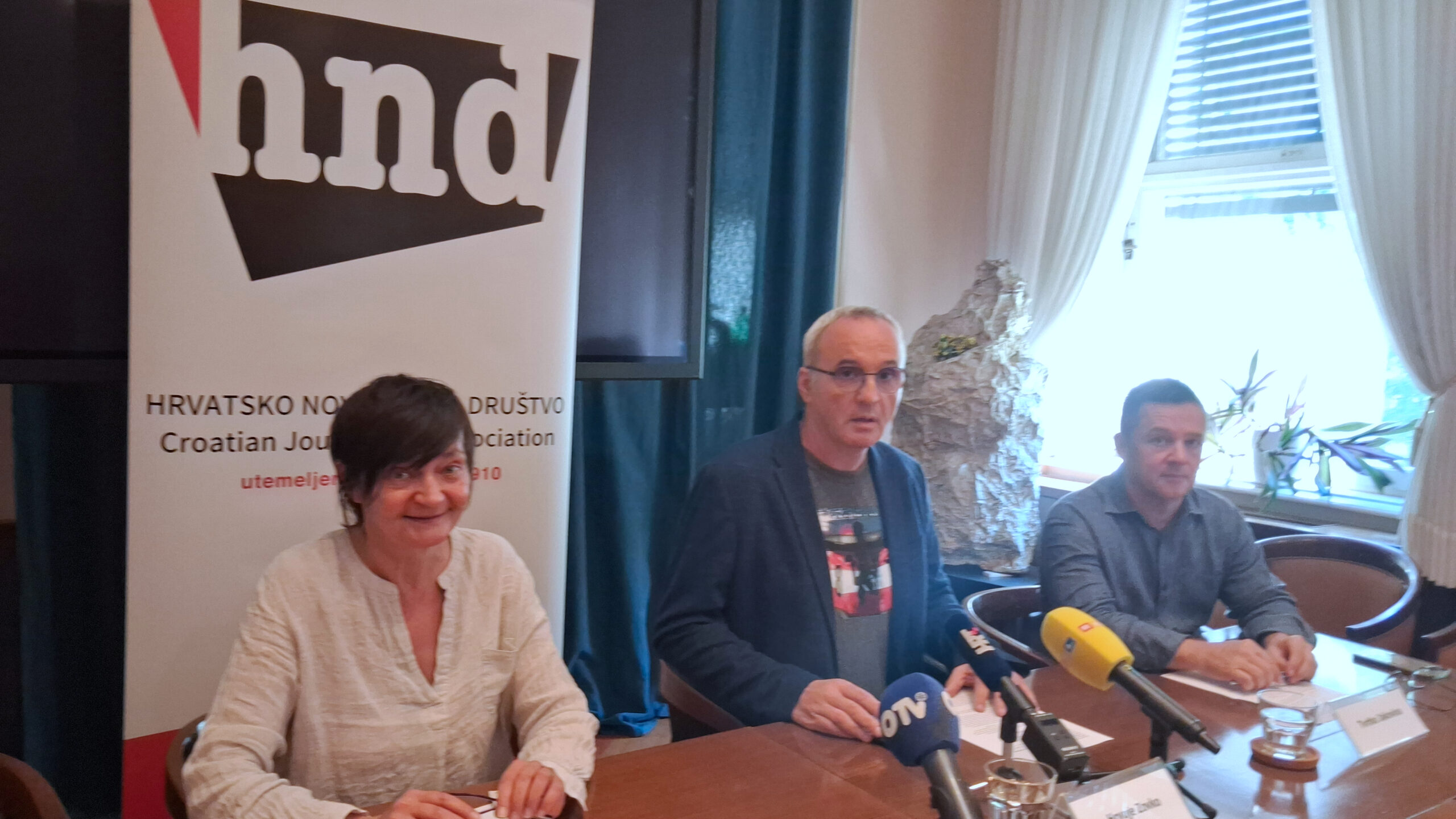Tri osobe sjede za stolom tijekom konferencije za medije u prostorijama Hrvatskog novinarskog društva. Ispred govornika nalaze se mikrofoni više medijskih kuća, a u pozadini je istaknut banner HND-a. Sudionici odgovaraju na pitanja novinara za okruglim konferencijskim stolom.